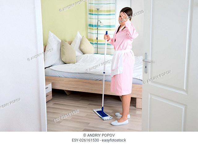 Exhausted Young Female Housekeeper With Mop In Hotel Room