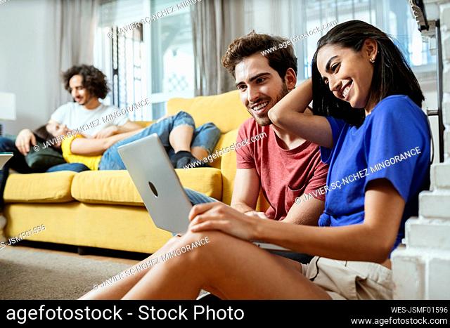 Smiling young couple sharing laptop while friends relaxing on sofa in living room at home