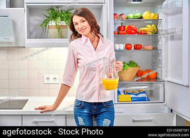 Front view of young attractive woman standing near opened refrigerator, keeping orange juice in hand, looking at camera and smiling
