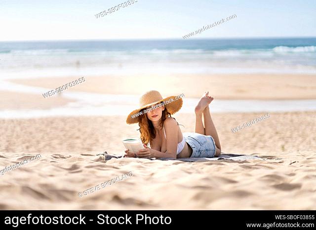 Young woman with book looking away while lying on sand at beach during vacations