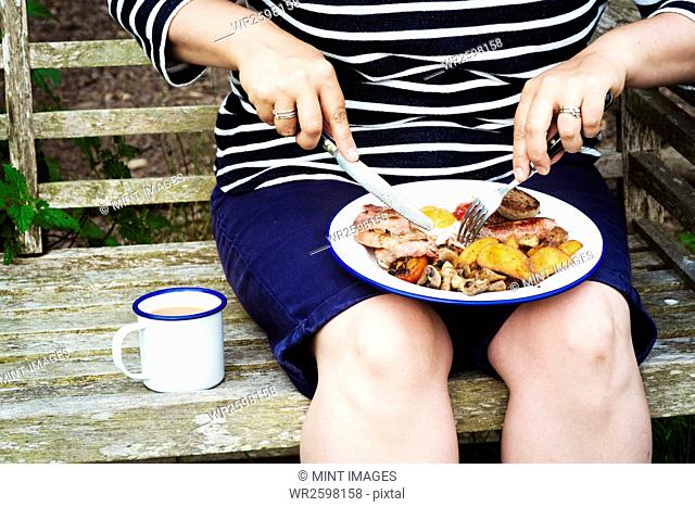 Woman sitting on a bench, eating from a plate of food balanced on her knees