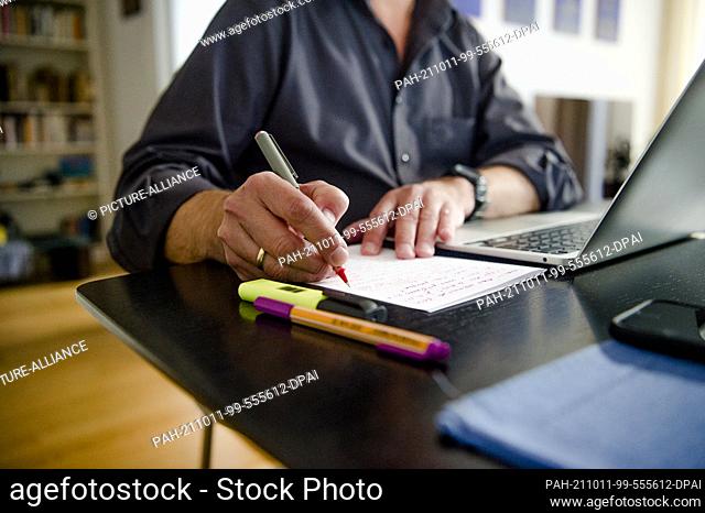 PRODUCTION - 01 October 2021, Bavaria, Munich: A man is sitting at a dining table at home. He is writing with a pen on a piece of paper next to a laptop