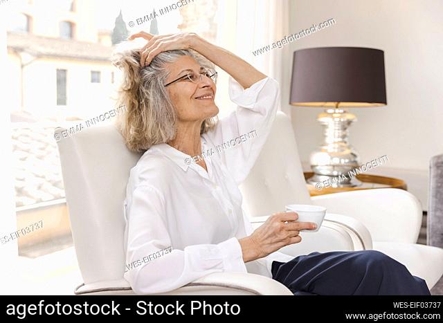 Smiling woman with hand in hair holding coffee cup sitting on armchair at home