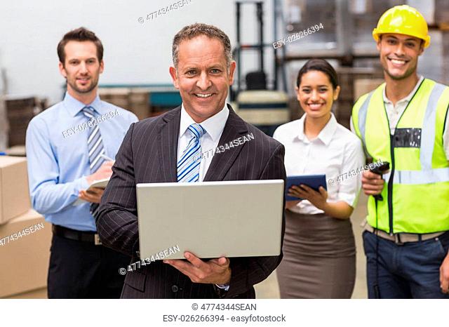 Smiling boss using laptop in front of his employees in a large warehouse