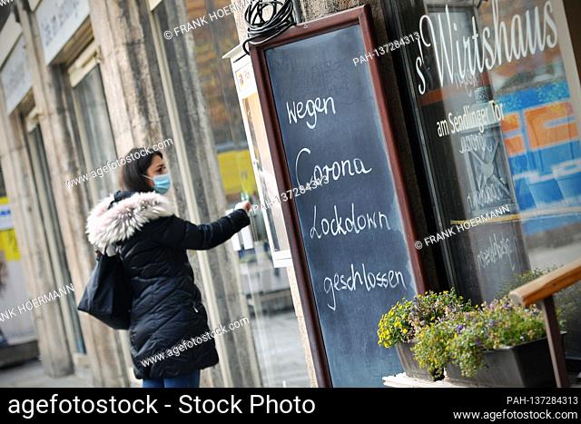 Topic picture: Coronavirus pandemic / consequences for gastronomy. A young woman stands with face mask, mask in front of a locked door of an inn next to a sign:...