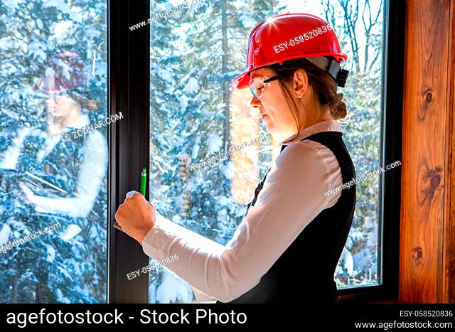 A caucasian woman inspector during house interior Inspection, she wearing a red hard hat and protective eyewear as she using a clipboard to take notes