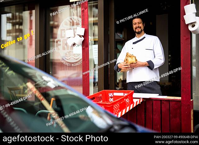 26 March 2020, Baden-Wuerttemberg, Weil am Rhein: Master baker Simon Fritz stands in the window of the improvised drive-in of his bakery and holds a loaf of...