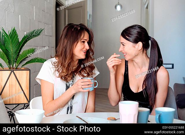Two woman friends breakfast in the kitchen and having fun, talking and laughing, sitting at the dining table at home