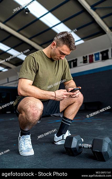 Male athlete using smart phone while crouching by dumbbell in gym