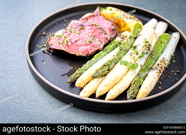 Traditional white and green asparagus with barbecue dry aged sliced beef fillet and fried potatoes served as closeup on a modern design plate