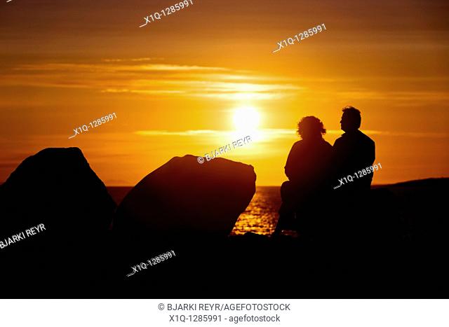 People sitting and enjoying the midnight sun setting over the Atlantic Ocean  Reykjavik Iceland