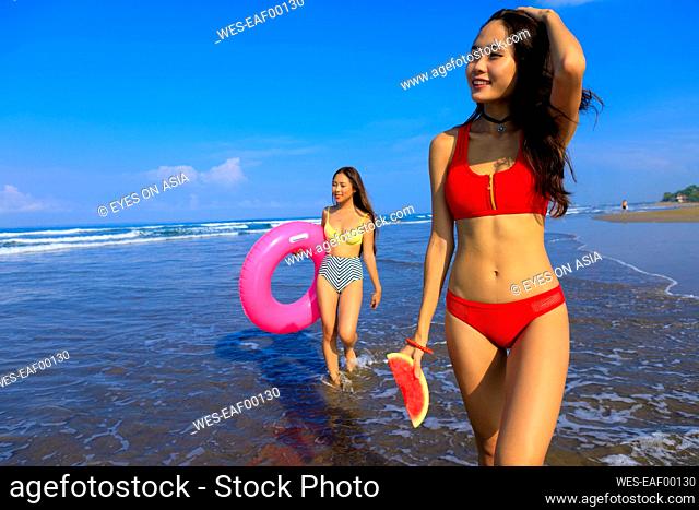 Smiling woman holding watermelon while female friend with swimming float walking through water on sunny day