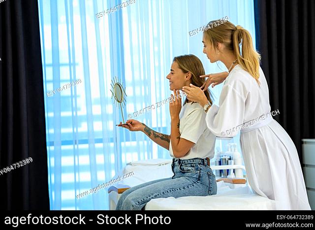 Beautiful woman sitting on couch with doctor cosmetologist, looking at mirror and examining skin after beauty skin treatment