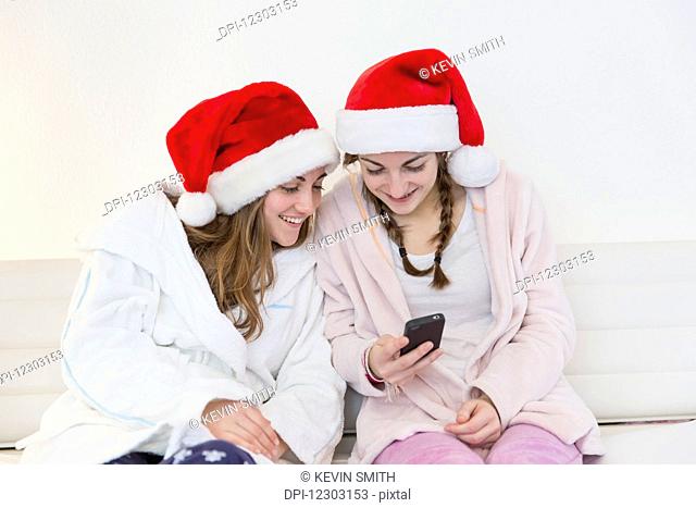 Two teenage sisters wearing santa hats, pajamas and robes and sitting together on a couch looking at a cell phone; Bonn, Nordrhein Westfalen, Germany