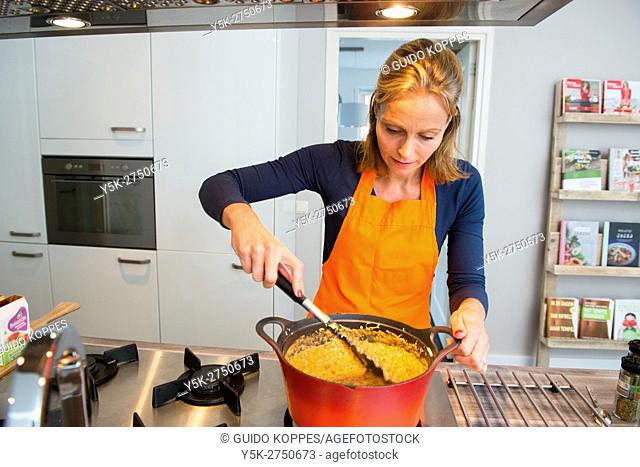 Kaatsheuvel, Netherlands. Mid adult woman stirring a red colored saucepan on her stove, busy making a Lasagna dish dinner