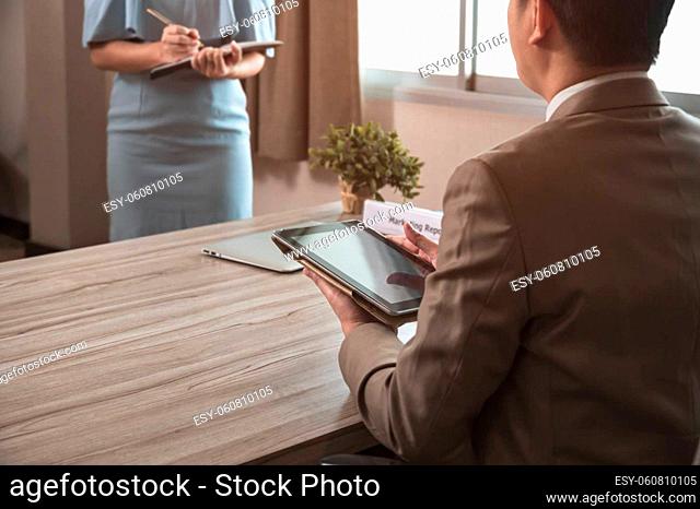 Businessman manager in brown suit using PC tablet while sitting at office desk and secretary in blue dress taking notes while standing in front of his boss