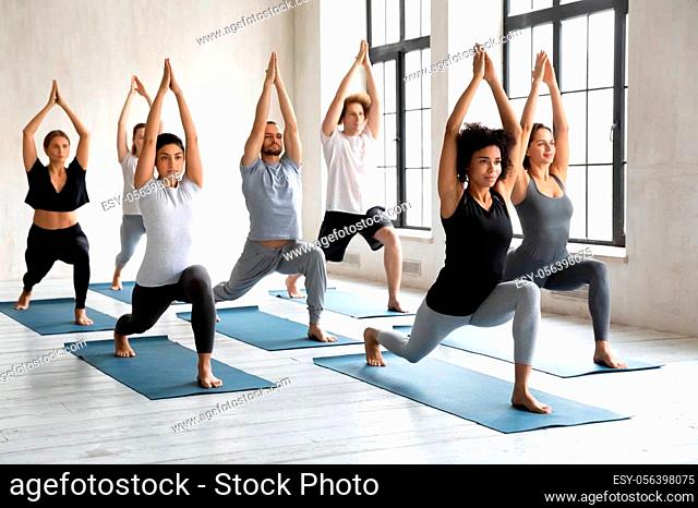 Young diverse young people with African American instructor doing Warrior one exercise, practicing yoga at group lesson, standing in Virabhadrasana pose