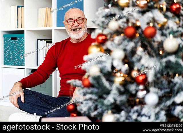 Smiling senior man wearing eyeglasses looking at Christmas tree sitting in living room