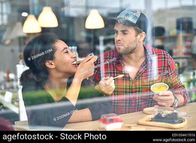 Black girl with her white boyfriend eating desserts in a cafe. She looks at him with a smile. Woman wears a dark longsleeve