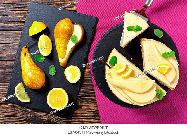 homemade pear sponge cake with cream cheese, decorated with mint, pear and lemon slices cut in slices on black slate plate, view from above
