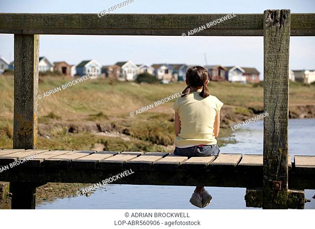 England, Dorset, Christchurch, A young girl sitting on a foot bridge looking towards beach huts in the distance at Christchurch in Dorset