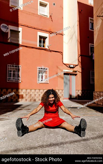 Smiling young woman wearing red dress and sitting on ground, stretching her legs