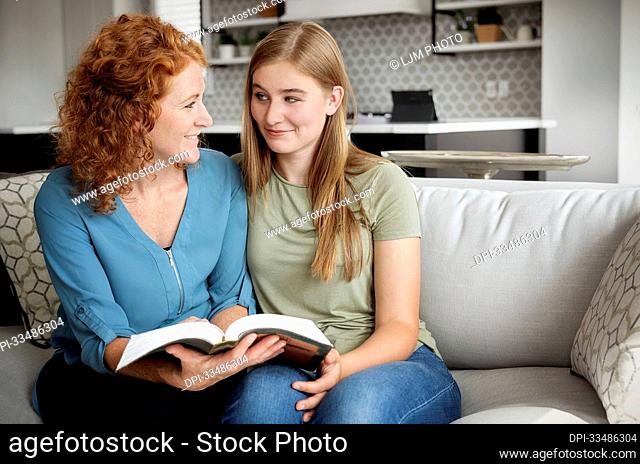Mother and teenage daughter sitting on a couch at home reading the Bible together; Edmonton, Alberta, Canada