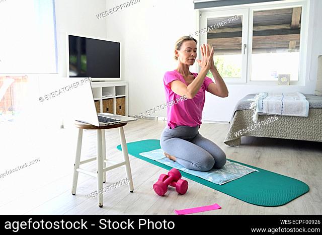 Mature woman with laptop practicing yoga on gym mat in living room