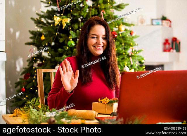The happy young woman is waving hand at laptop camera. Woman sitting in front of a laptop on the background of the Christmas interior