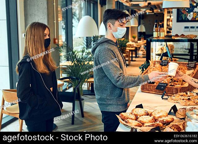 People standing in a queue to buy coffee and pastry in the coffee shop to go. Man taking coffee cup from barista. People standing at counter wearing the face...