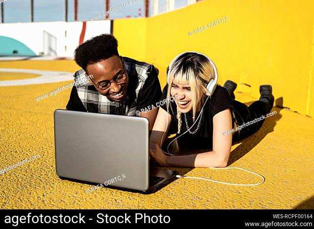 Laughing young couple using headphones and laptop outdoors having fun