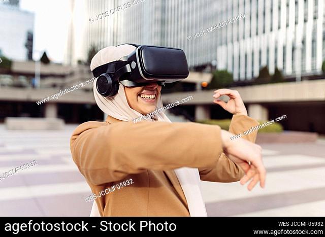 Happy young businesswoman wearing virtual reality simulator gesturing at office park on sunny day