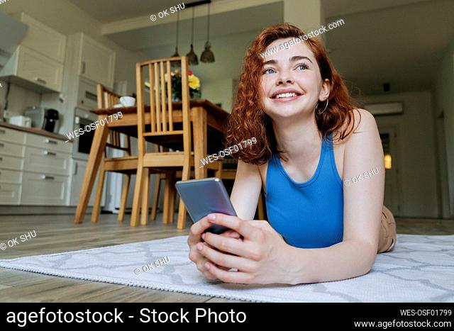 Happy young woman with smart phone lying on rug at home