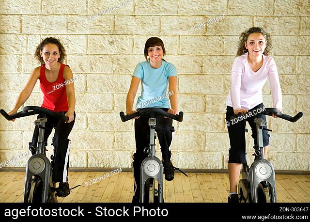 Three happy, young women working out on exercise bicycle at the gym. Front view, looking at camera
