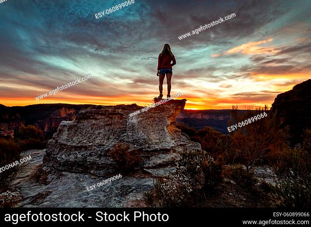 Female hiker stands on mountain peak rocky outcrop along the cliifs of Blue Mountains, taking in spectacular views as the sun sets