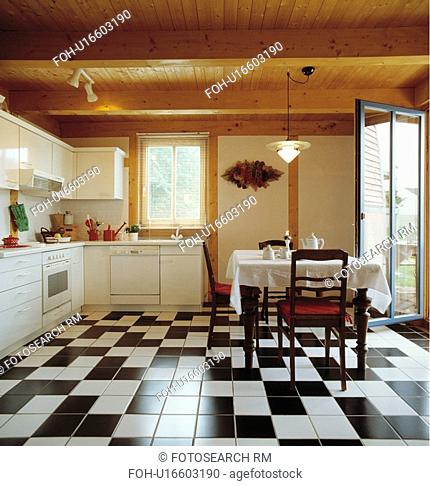 Wooden ceiling and black+white ceramic tiles floor in modern white country kitchen with traditional table and chairs