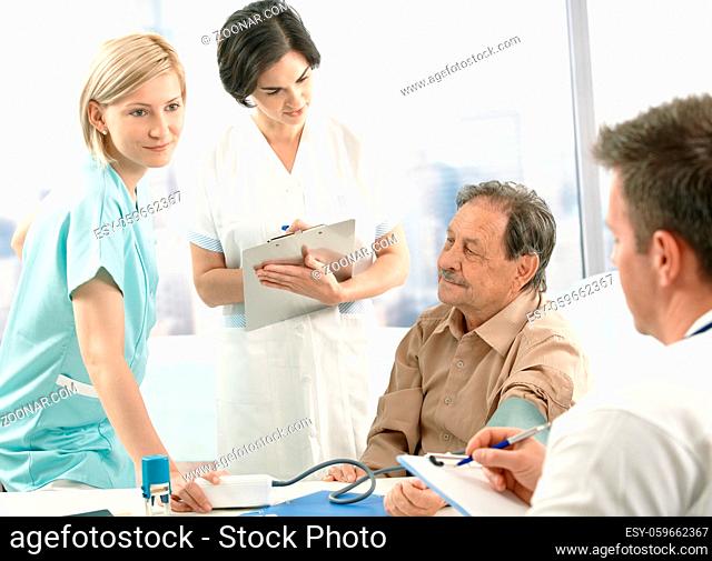 Medical team measuring blood pressure of senior patient, nurse assisting, doctors taking notes on clipboard