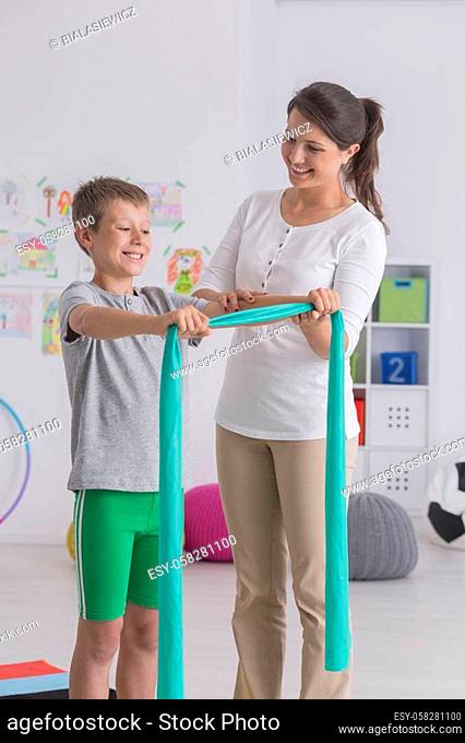 Boy doing exercise with elastic band during meeting with pretty physiotherapist