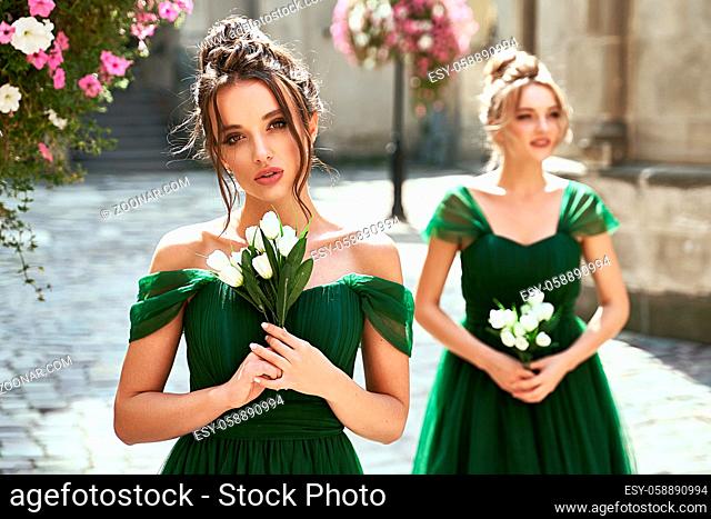 Two beautiful bridesmaids girls blonde and brunette ladies wearing elegant full length off-the-shoulder green chiffon bridesmaid dress and holding flower...