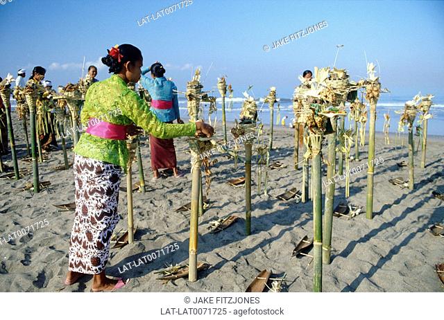 Bamboo poles and offerings on beach. Religious ritual. Offerings to gods. View out to sea. Sand. People in traditional dress. Women in sarongs