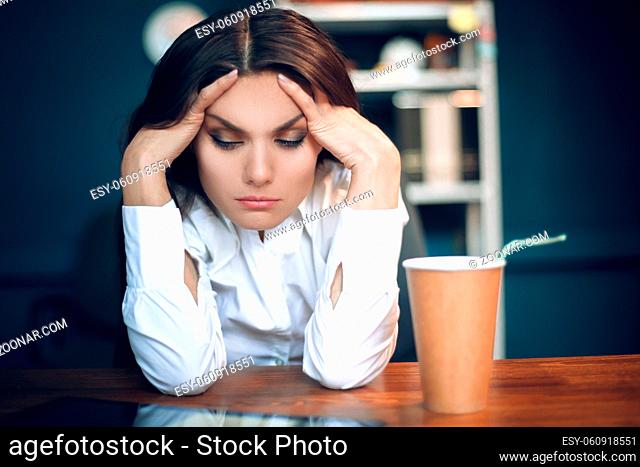 Tired gorgeous girl with her head in her hands. Lady wearing white blouse sitting alone with her eyes closed at wooden table with brown coffee cup by her side