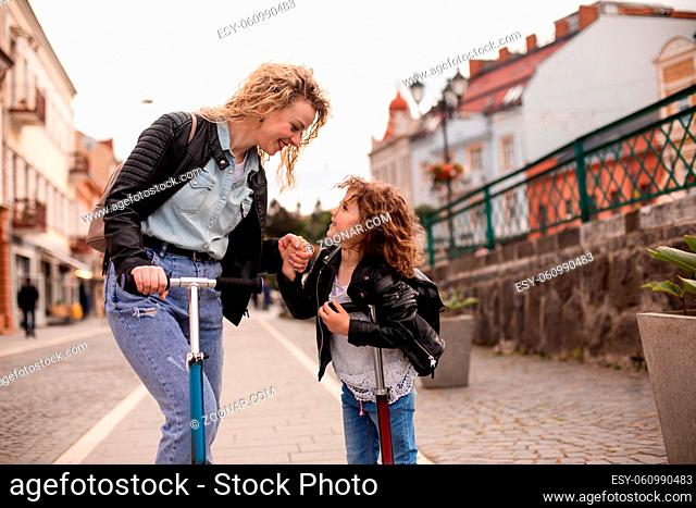 The young woman with a little girl are standing with scooters in the city, look each other