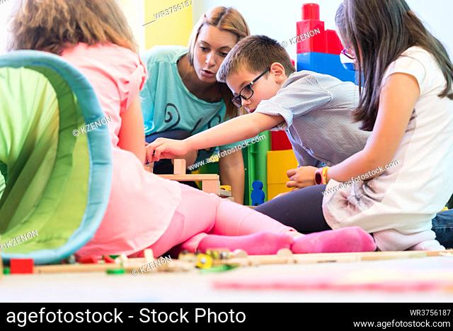 Cute pre-school boy cooperating with his colleagues at the construction of a structure, made of wooden toy blocks under the guidance of a young kindergarten...