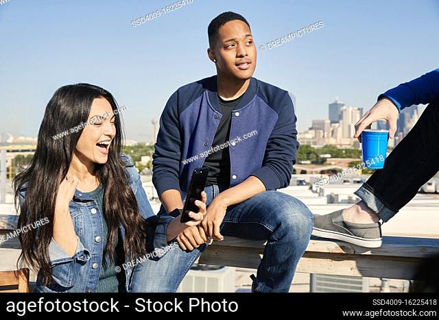 Group of young co-workers hanging out on rooftop patio laughing and having a drink, woman looking at mobile and guy looking off camera