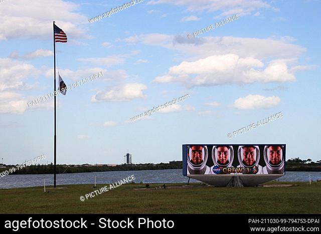 29 October 2021, US, Cape Canaveral: A US flag flies next to a video screen with the faces of the four astronauts of Crew-3 (l-r): Raja Chari, Kayla Barron