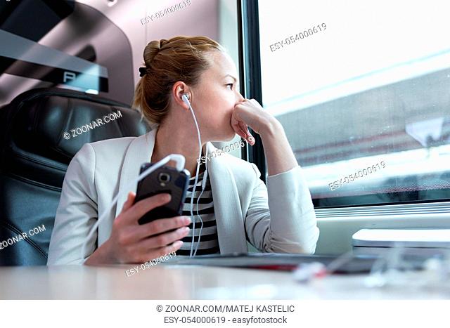 Thoughtful businesswoman looking trough the window, listening to podcast on cellphone using headphone set while traveling by train in business class seat