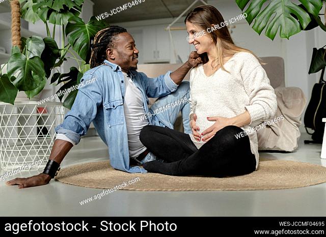 Smiling man looking at pregnant woman touching belly on rug in living room