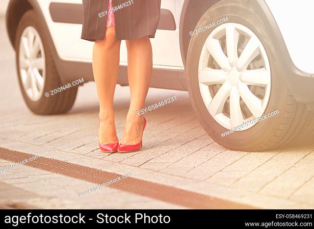 Close-up picture of low view of woman#39;s legs near white car. Professional model standing on fashionable expensive pink high heels. Toned image