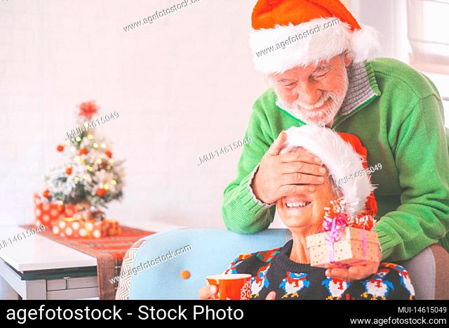 Senior couple in warm clothing and santa hat, man covering eyes of old wife with a surprise gift for her. Loving old romantic heterosexual couple celebrating...