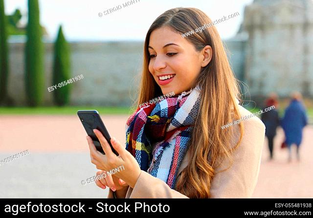 Happy cheerful young woman typing with finger on smart phone on city square wearing coat and scarf in winter